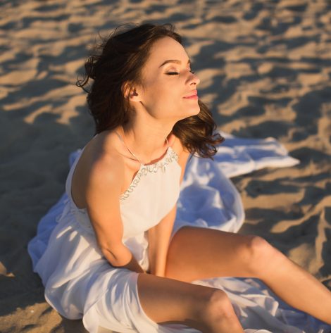 Beautiful girl bride in a white dress sitting on beach sand at sunset